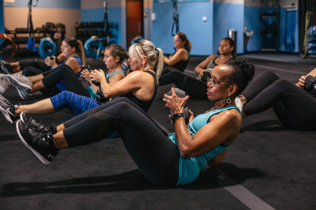 Fitness class with a group of women in gym sitting down on floor with weights with feet raised off floor