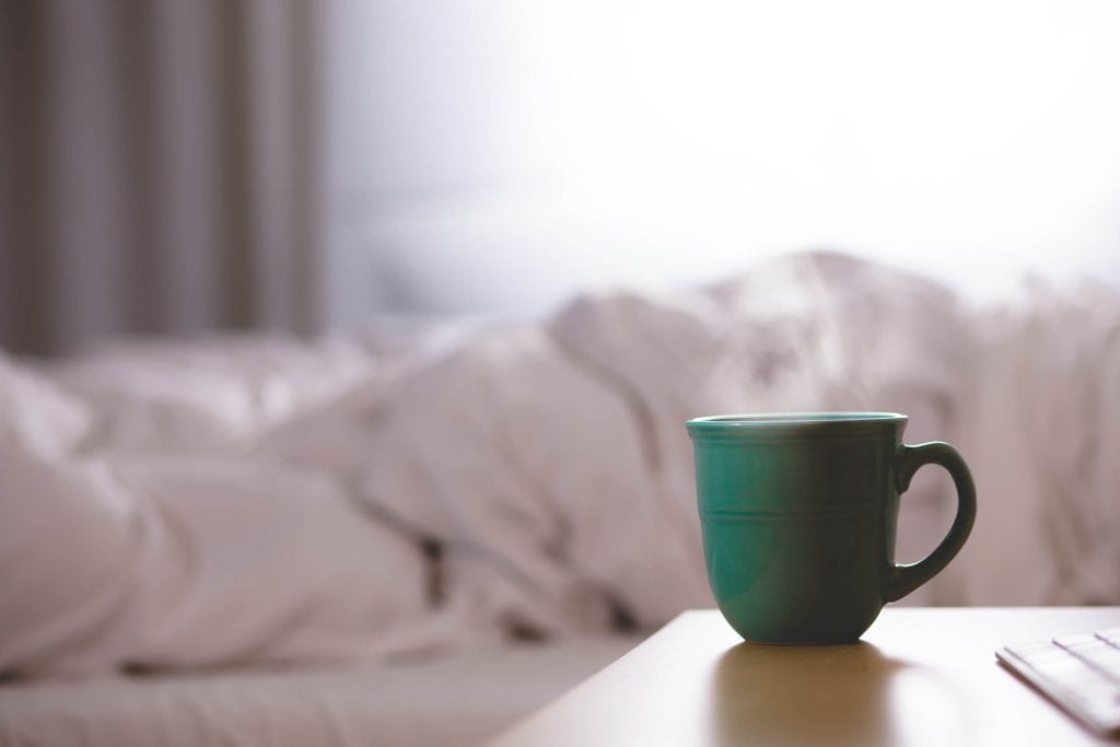 Green cup of coffee on edge of table next to unmaid paid in bedroom