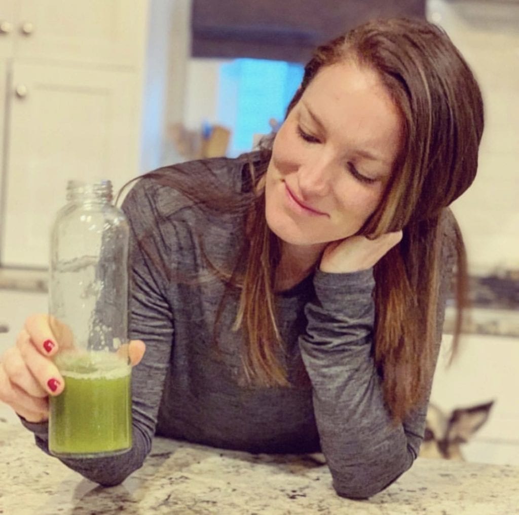 Woman in grey long sleeve t shirt leaning on countertop holding a clear bottle of celery juice.