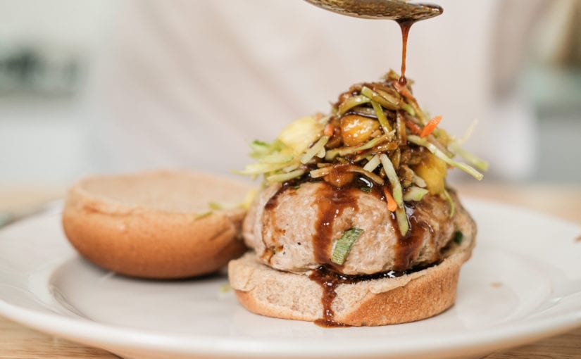 Spoon drizzling dark sauce over turkey burgers with asian salad on top sitting on white glass plate on table
