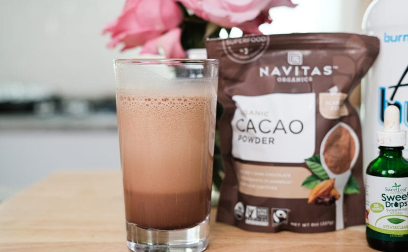 Clear glass filled with chocolate protein drink sitting on countertop with bag of cocoa powder