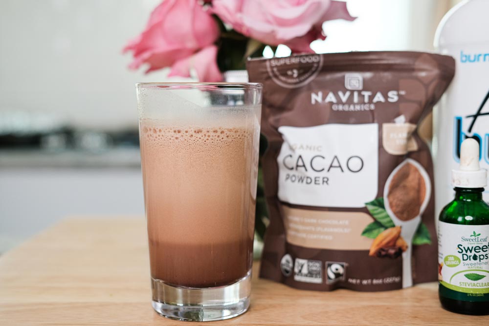 Clear glass filled with chocolate protein drink sitting on countertop with bag of cocoa powder