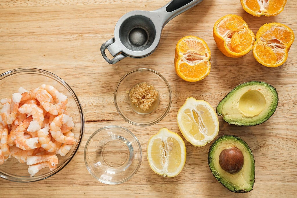 Clear glass bowl of shrimp with a grey juicer with sliced lemons, oranges and avocados on table with small glass bowl of water and garlic.